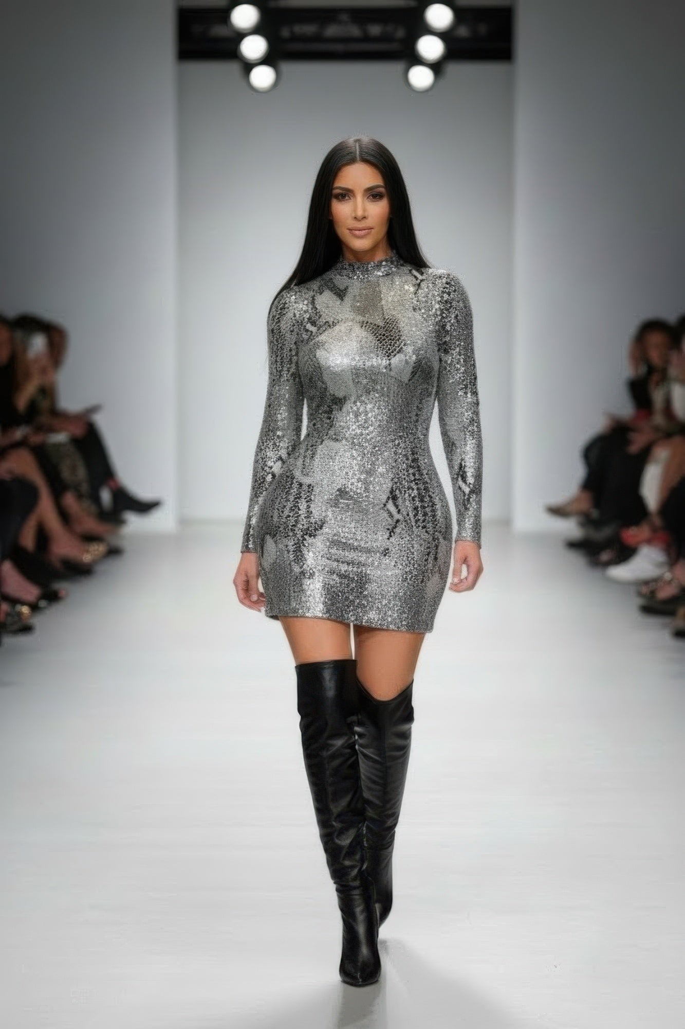 Model walking on a runway wearing a silver sequin dress and black knee-high boots