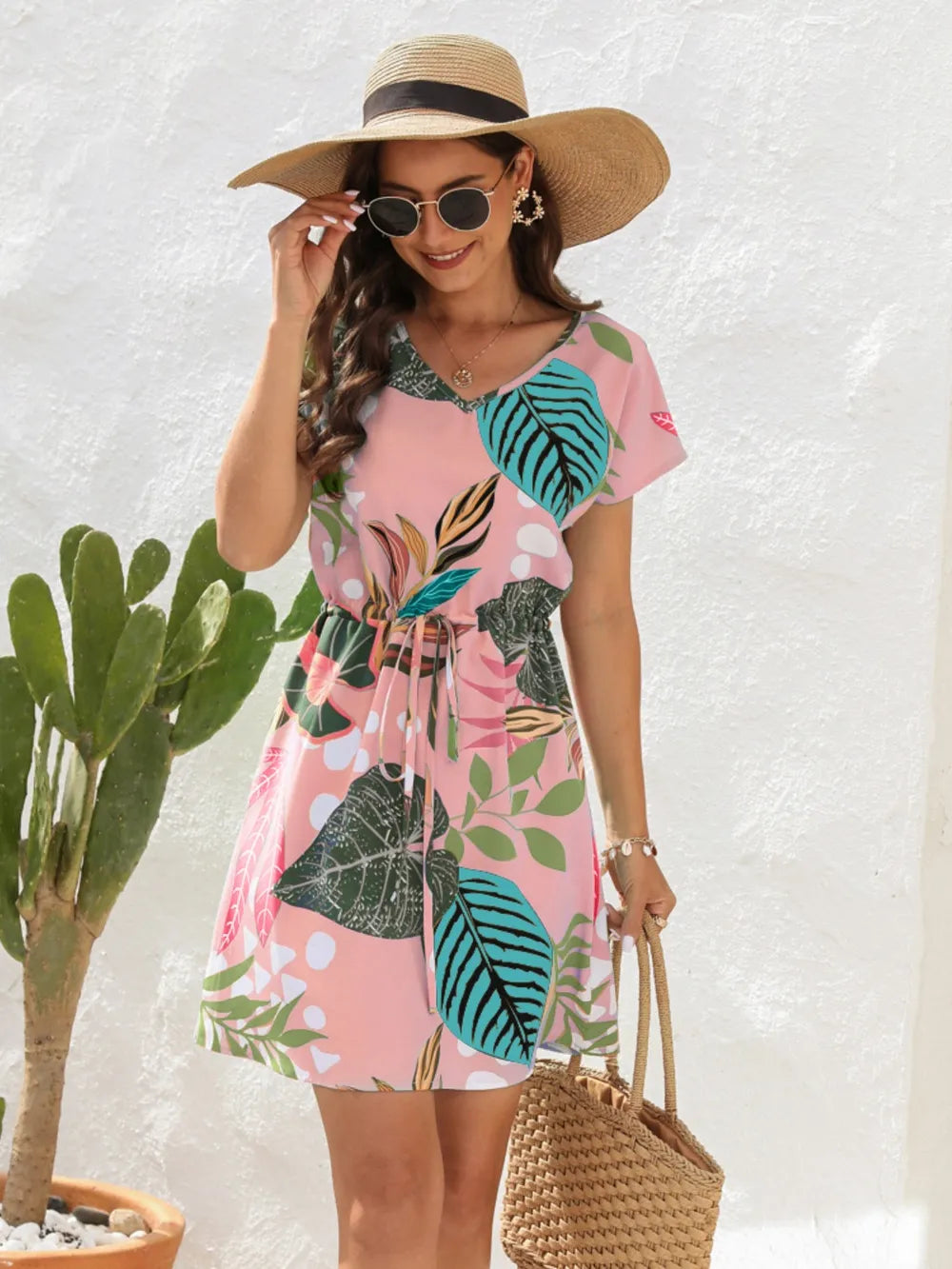 Woman wearing a pink dress with leaf pattern, straw hat, and sunglasses, standing next to a cactus plant.