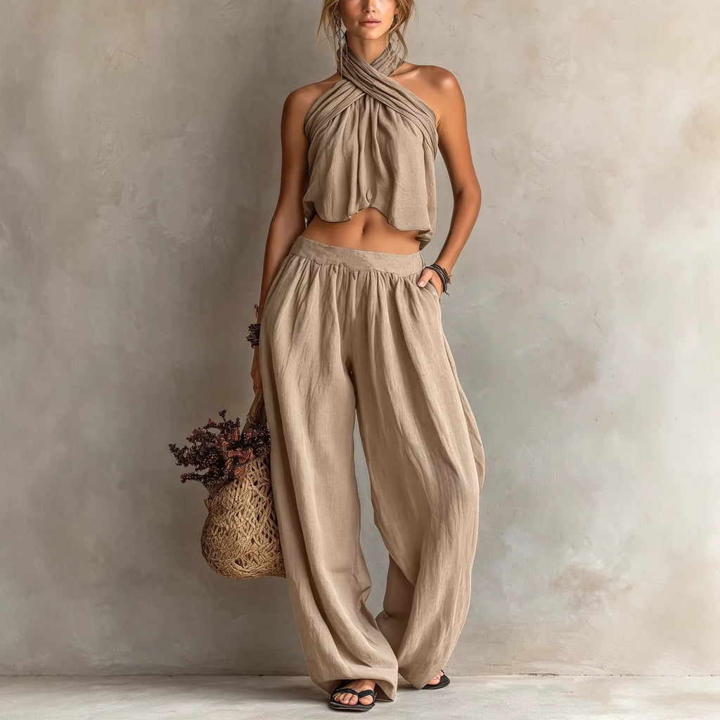 Woman wearing a beige halter top and wide-leg pants, holding a woven bag against a neutral background.